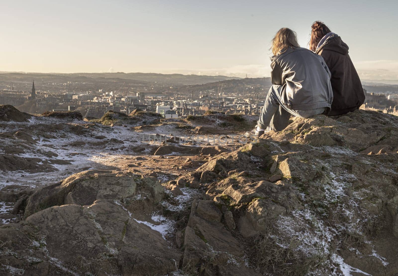 Edinburgh, Scotland - Jan 17, 2024 - Two young women sitting relaxing on top of rock over the mountain and enjoying looking at scenic view of cityscape. Two female Sitting down enjoying the amazing view from the mountain top of Salisbury Crags, Destinations in Europe, Space for text, Selective focus.