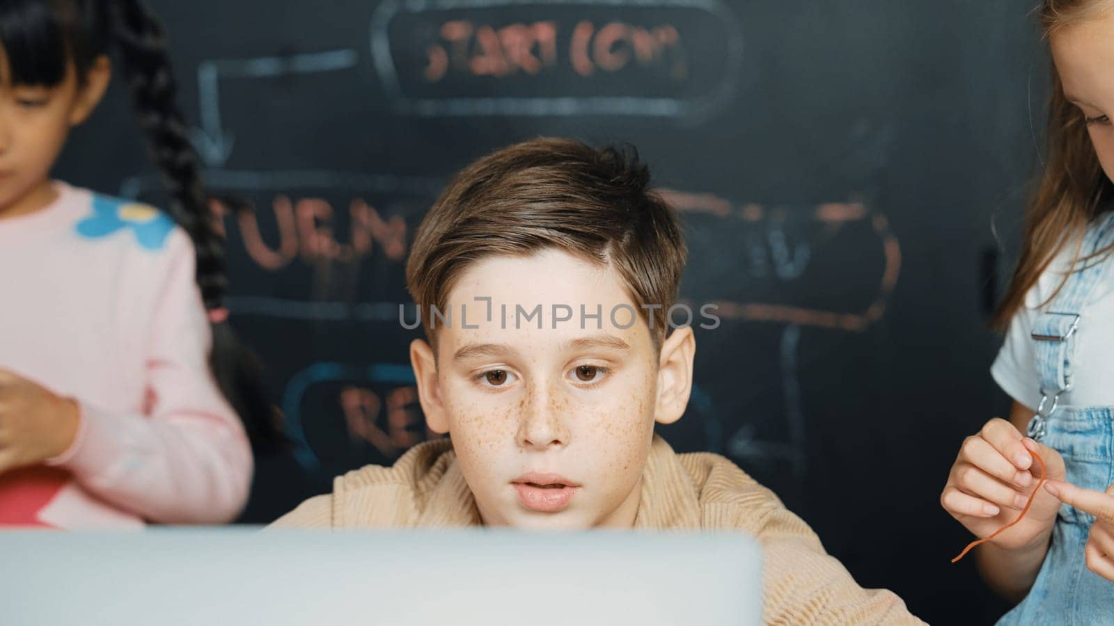 Closeup of boy using laptop programing engineering code and writing program while group of diverse kid holding controller in STEM technology classroom at blackboard written with prompt. Erudition.