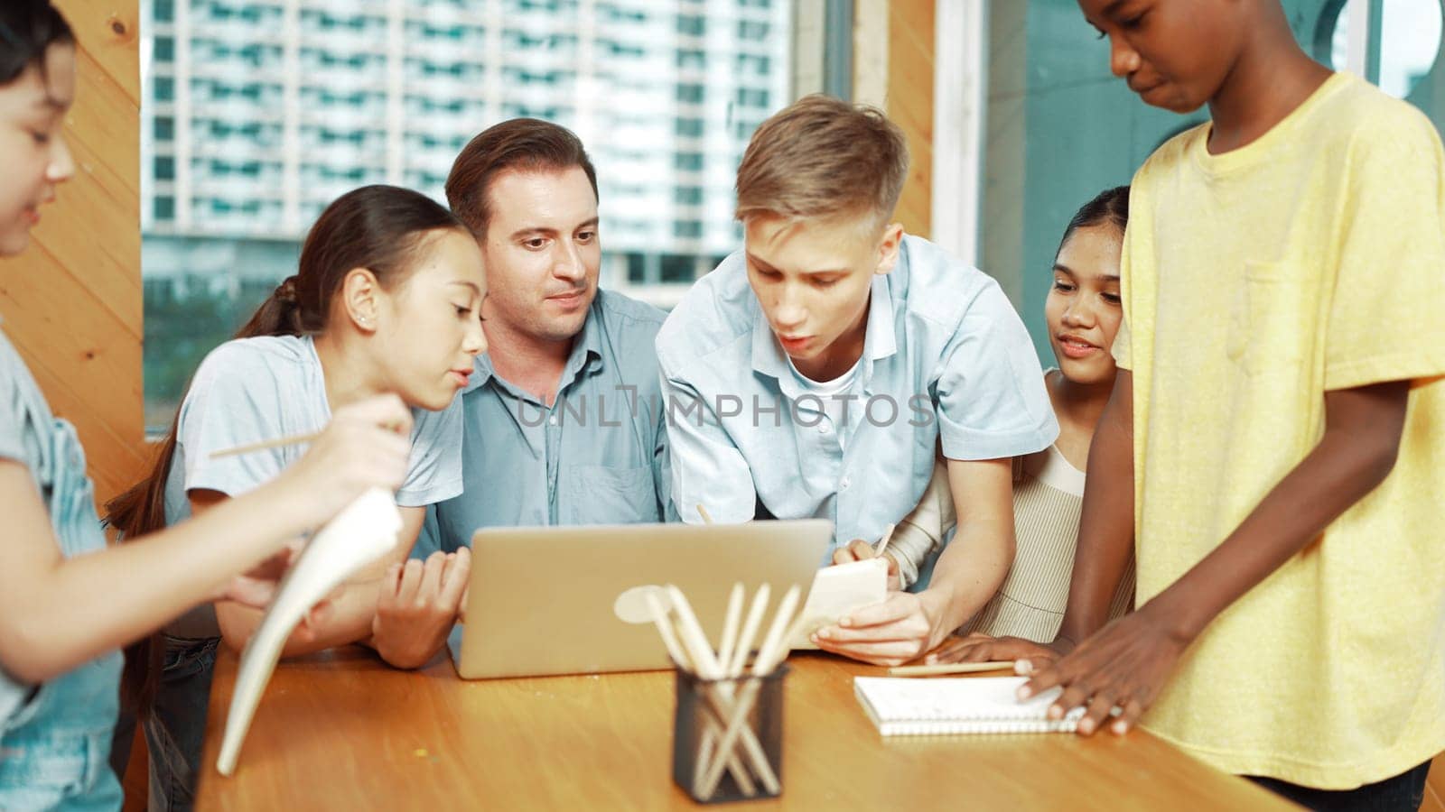 Caucasian teacher giving advise to young student while boy lecture comment. Professional instructor write comment surrounded by multicultural children at table with laptop and paper. Edification.