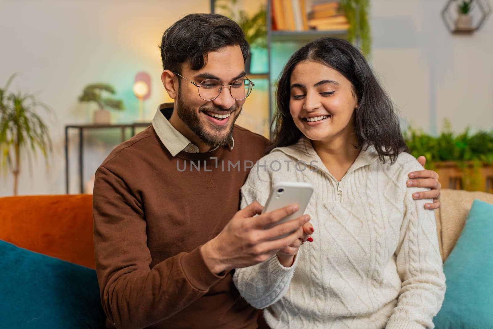 Multiracial excited couple sitting on sofa and swipe smartphone at home. Happy Indian man and woman discussing together spend time, relaxing and enjoying communicate on mobile phone in room apartment