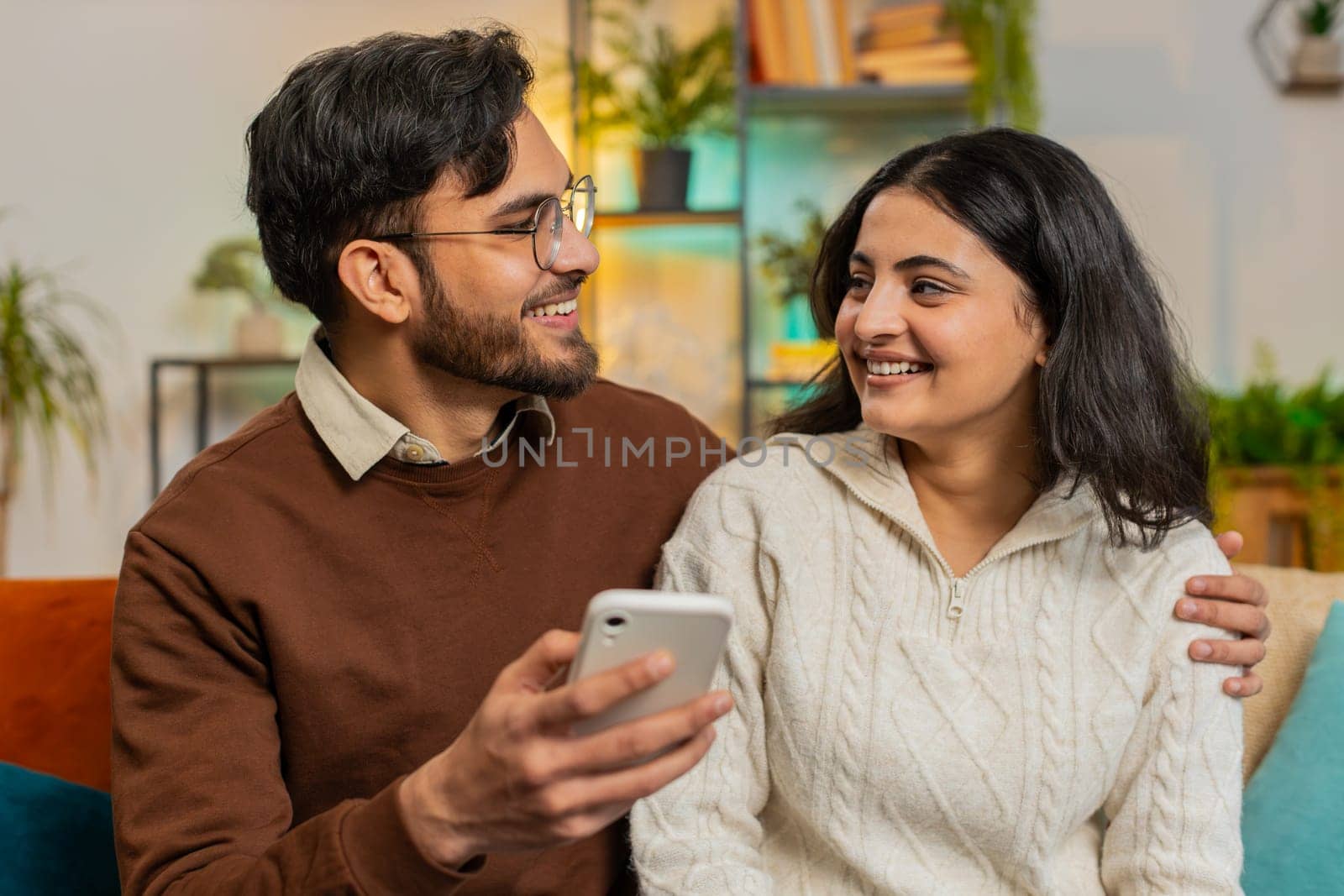 Multiracial young couple sitting on sofa and swipe smartphone at home. Happy Hispanic man and woman discussing together spend time, relaxing and enjoying communicate on mobile phone in room apartment