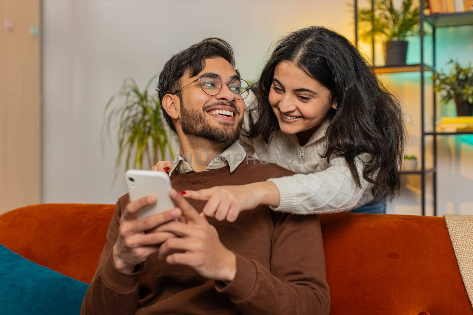 Smiling Arabian woman talking with boyfriend swiping smartphone sitting on sofa at home. Multiethnic young couple using mobile phone together making order online delivery in living room apartment.