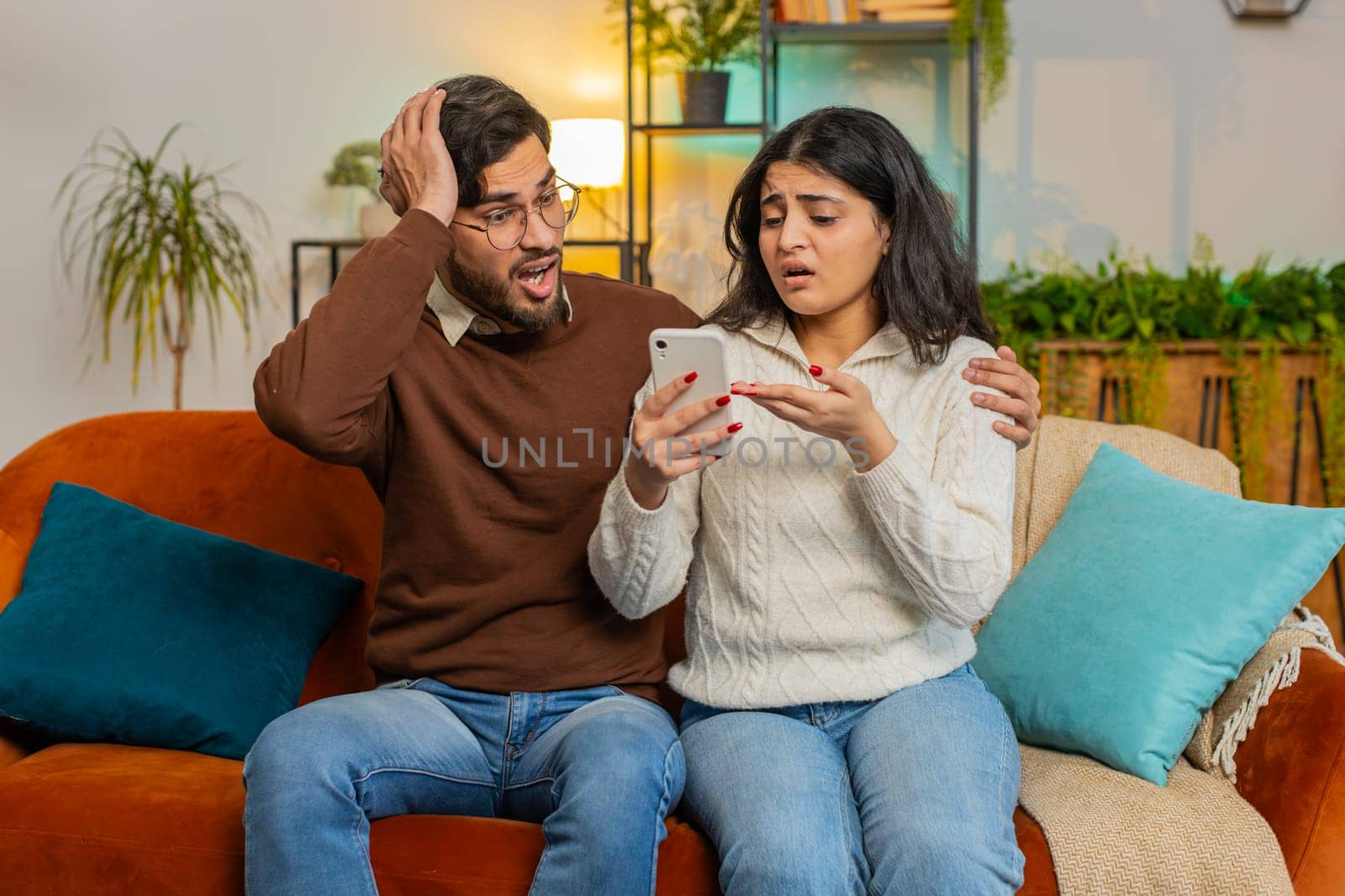Disappointed multiethnic couple man woman reading together bad news on smartphone at home on sofa by efuror