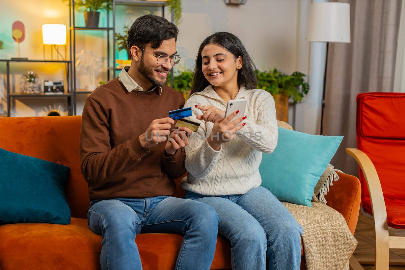 Happy young couple together doing online shopping by entering credit card number on smartphone while sitting on sofa at home. Smiling family are satisfied with a good promotion on goods in apartment