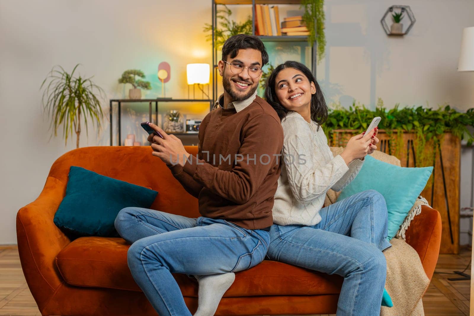 Side view of cheerful diverse couple sitting back to back using smartphones talking on home sofa by efuror