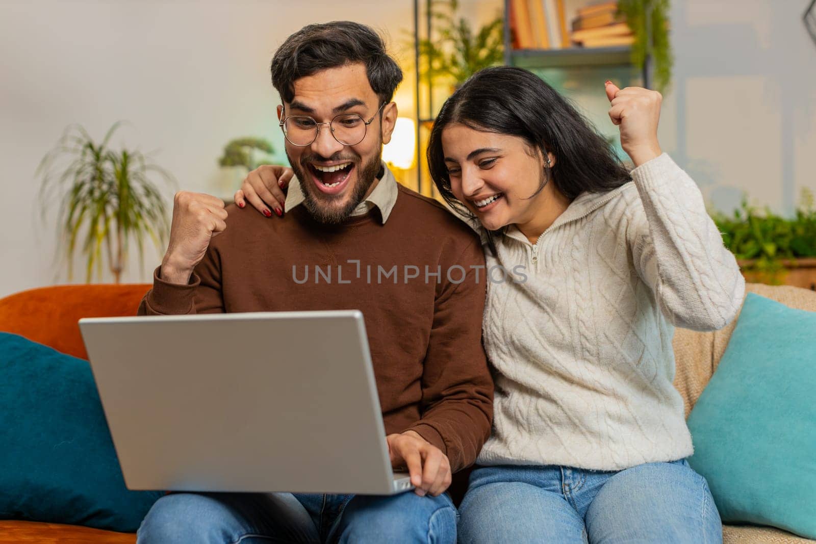 Cheerful young diverse couple in casual clothes using laptop surprised with email winning lottery online game jackpot on sofa in living room at home. Happy Hispanic family clenching fists in apartment