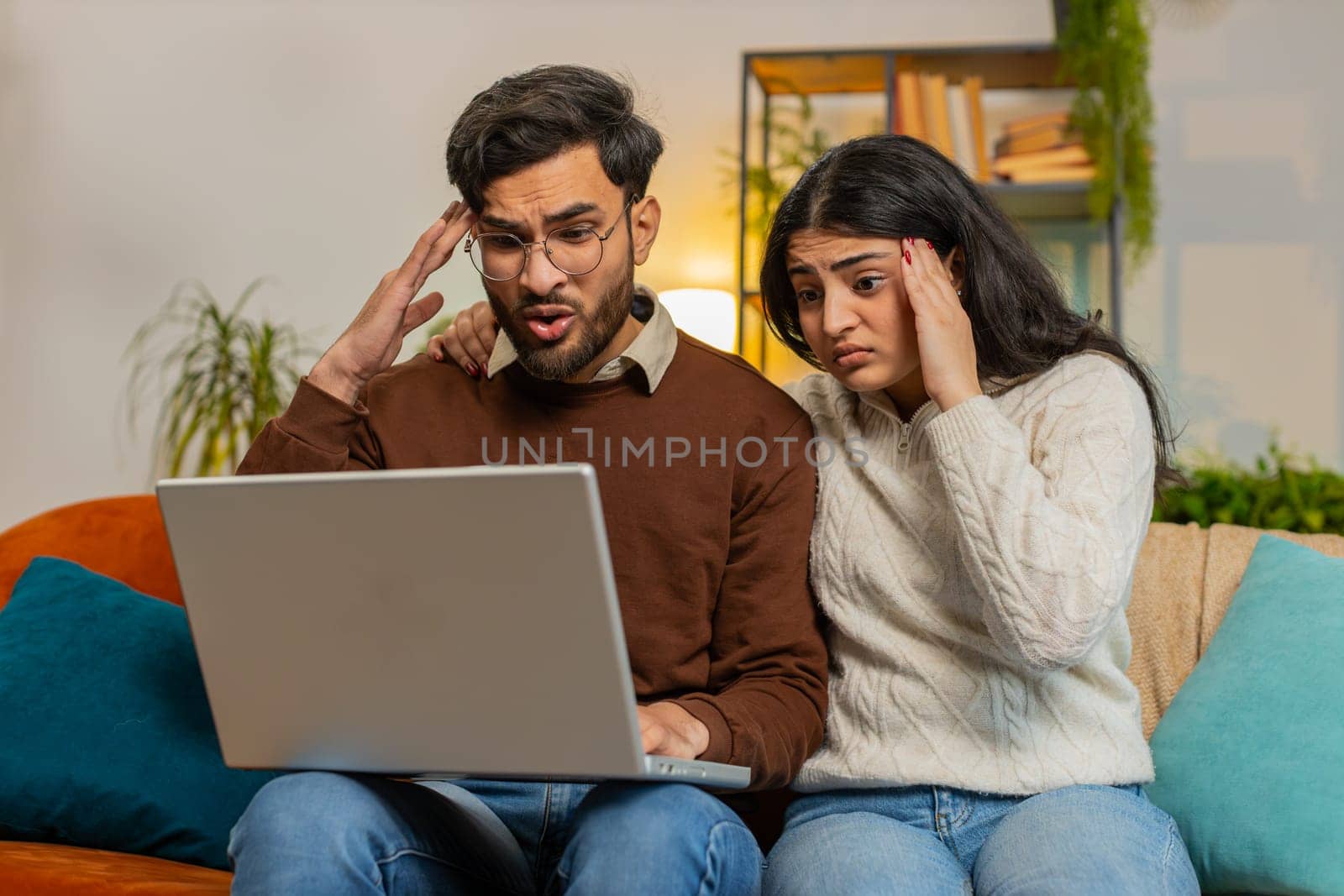 Shocked young diverse couple using laptop and covering mouth sitting on sofa in room at home. Upset disappointed Hispanic family man woman together reading bad news fail lose on netbook in apartment.