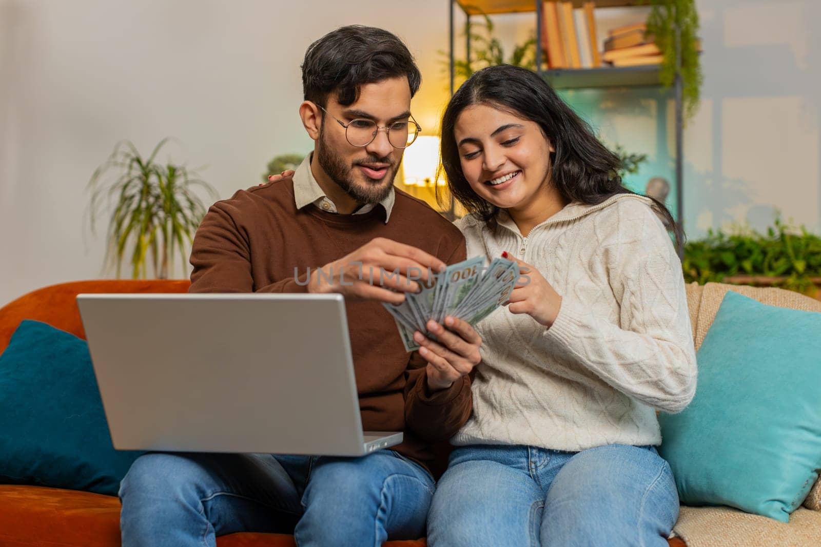 Happy young Indian couple counting money and booking tickets for holiday vacation sits on sofa in living room at home. Diverse family holding banknotes planning online investment budget home finance