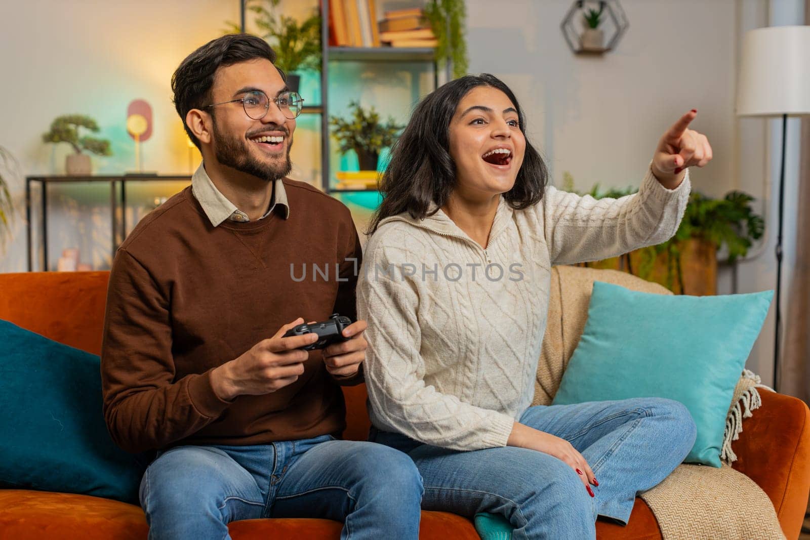 Cheerful young Indian couple using joystick controller playing video game fun enjoying sitting on sofa in living room. Diverse family enjoying success winning online game during weekend in house