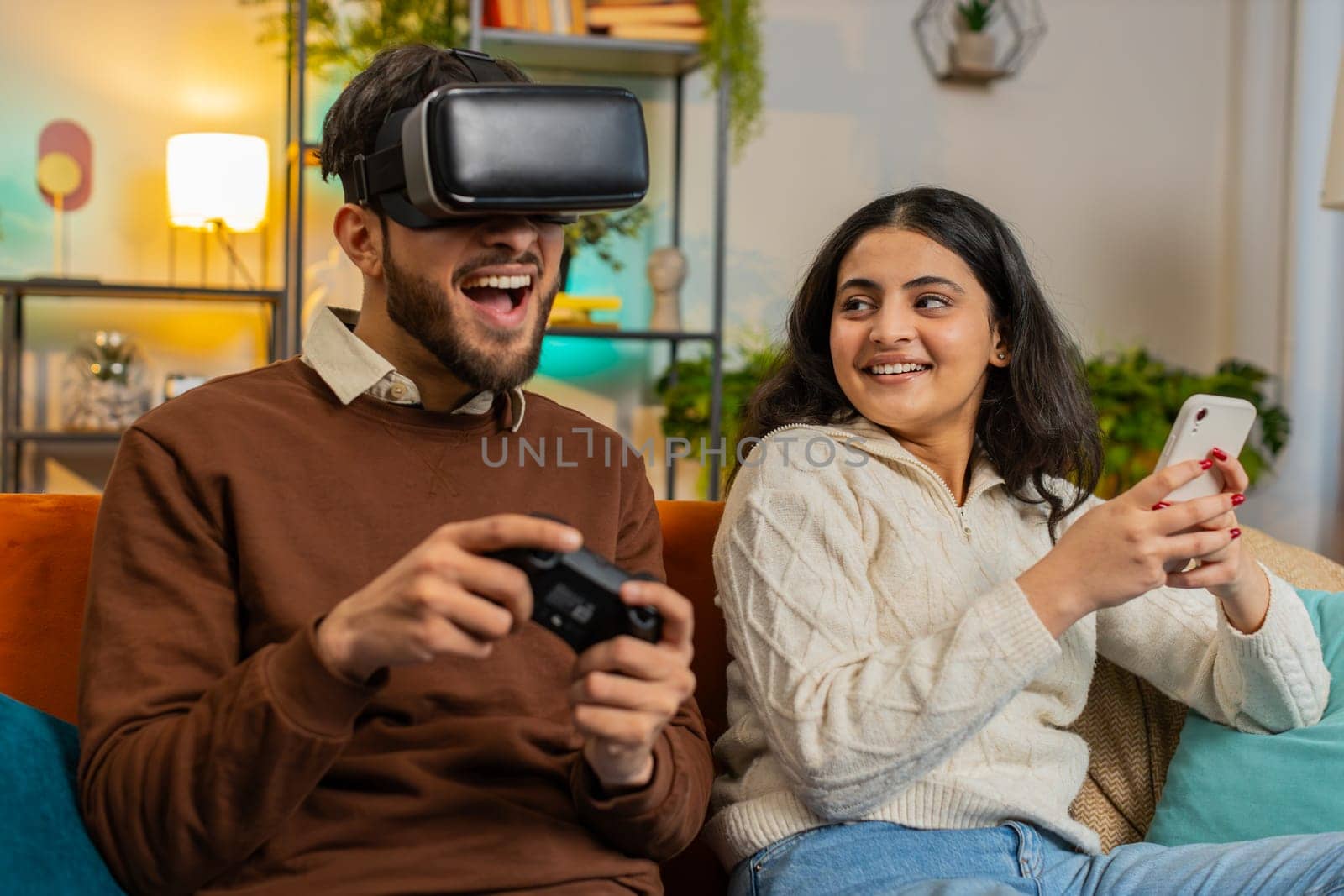 Happy exited man wearing VR goggles playing videogame with joystick sitting beside Indian girlfriend using smartphone sitting on sofa in living room at home. Happy family spending weekend in apartment