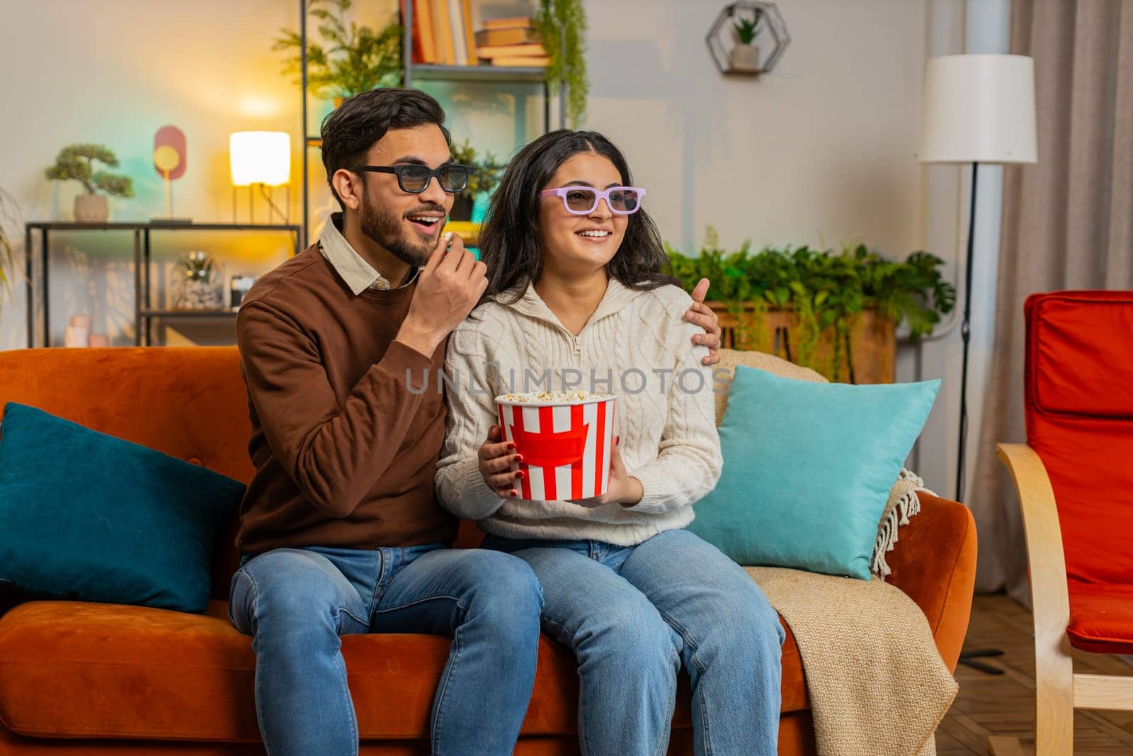 Cheerful young Arabian couple in 3D glasses eating popcorn and watching comedy movie sitting on sofa in living room at home. Smiling diverse family laughing enjoying film during weekend in apartment.