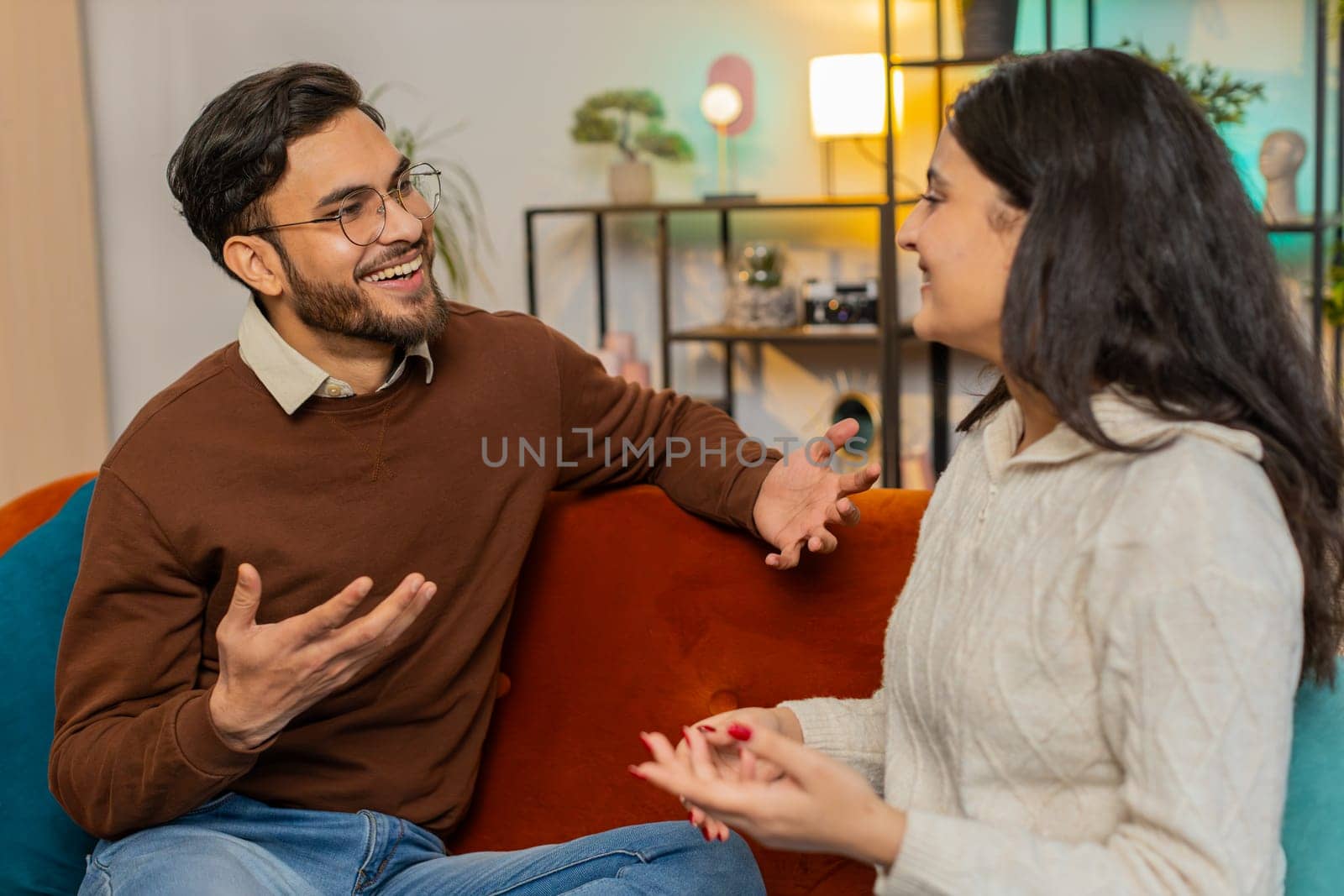 Smiling young family couple in casual clothes sitting on sofa together having lovely conversation in living room at home. Hispanic man discussing with Indian girlfriend relaxing on couch in apartment.