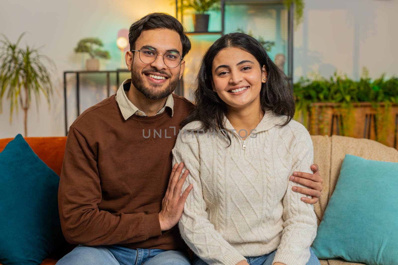 Loving young diverse couple looking at camera and smiling sitting arm around relaxing on sofa. Happy Indian family spending leisure time together on couch looking at camera in living room at home.