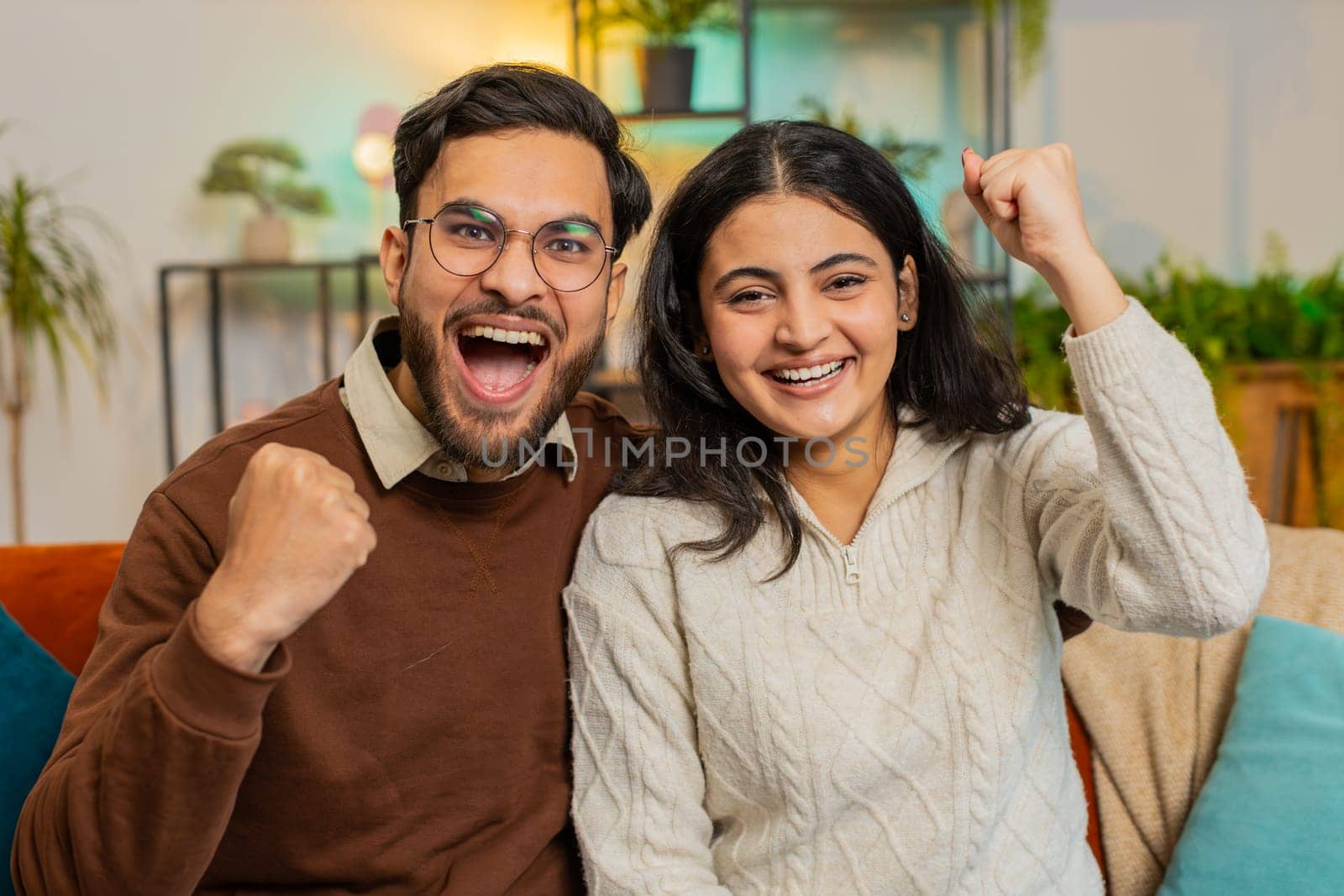 Indian couple celebrating success, clenching fists, showing thumbs up and giving high-five at home by efuror