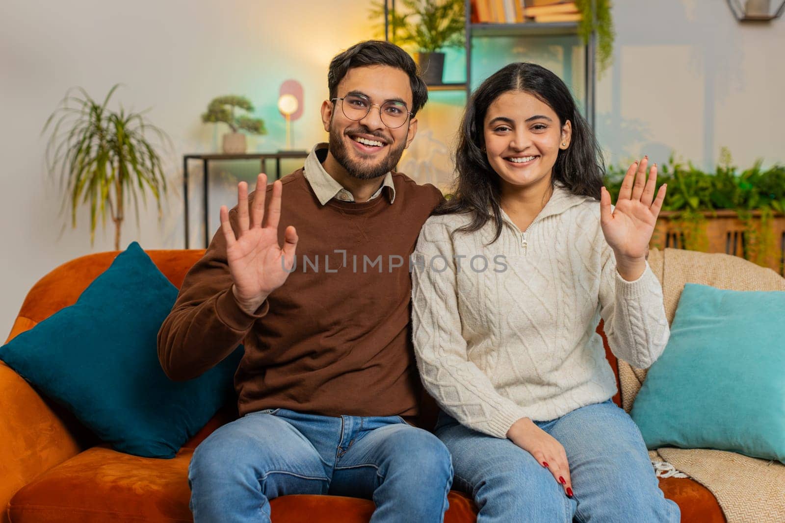 Portrait of smiling young diverse couple smiling at camera and waving gesturing hello, hi, greeting or goodbye. Relaxed family sitting arm around relaxing together on couch in living room at home.