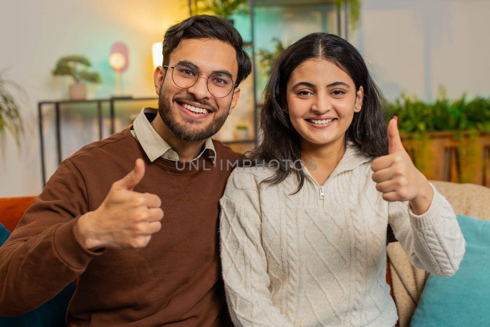Smiling young Hispanic couple sits arm around show thumbs up on sofa in living room at home. Portrait of happy diverse family in casual clothes together looking at camera giving approval good feedback