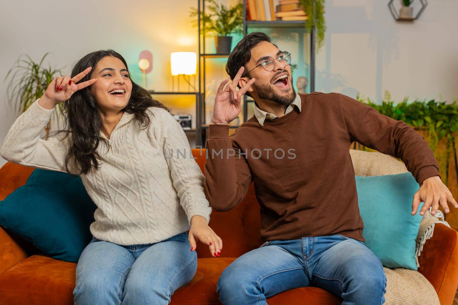 Cheerful excited young Hispanic couple dancing giving high five sitting on sofa in living room at home. Smiling happy diverse girlfriend and boyfriend enjoying weekend together on couch in apartment.