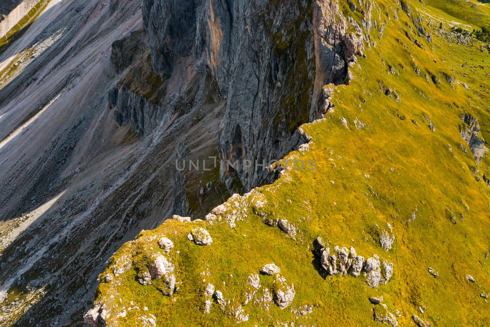 Green grass grows on steep slope of rocky Seceda mountain on sunny summer day. Rocky ridgeline in touristic Italian Alps aerial view
