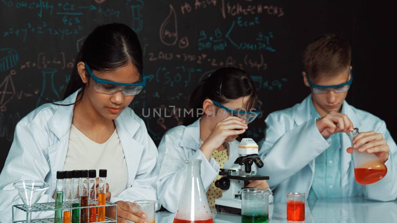 Smart teenager drop sample in microscope while girl inspect carefully at blackboard with chemistry theory. Professional scientist pour chemical product in tube and prepare for experiment. Edification.