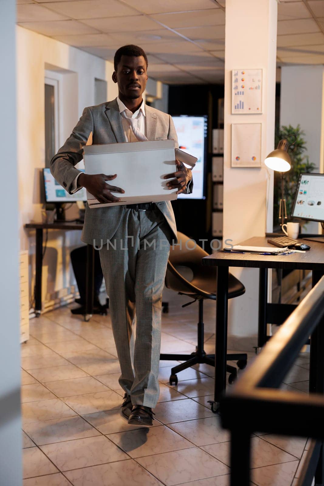 Sad manager carrying cardboard box with belongings after being dismissed from startup office. African american businessman leaving office late at night. Business management concept