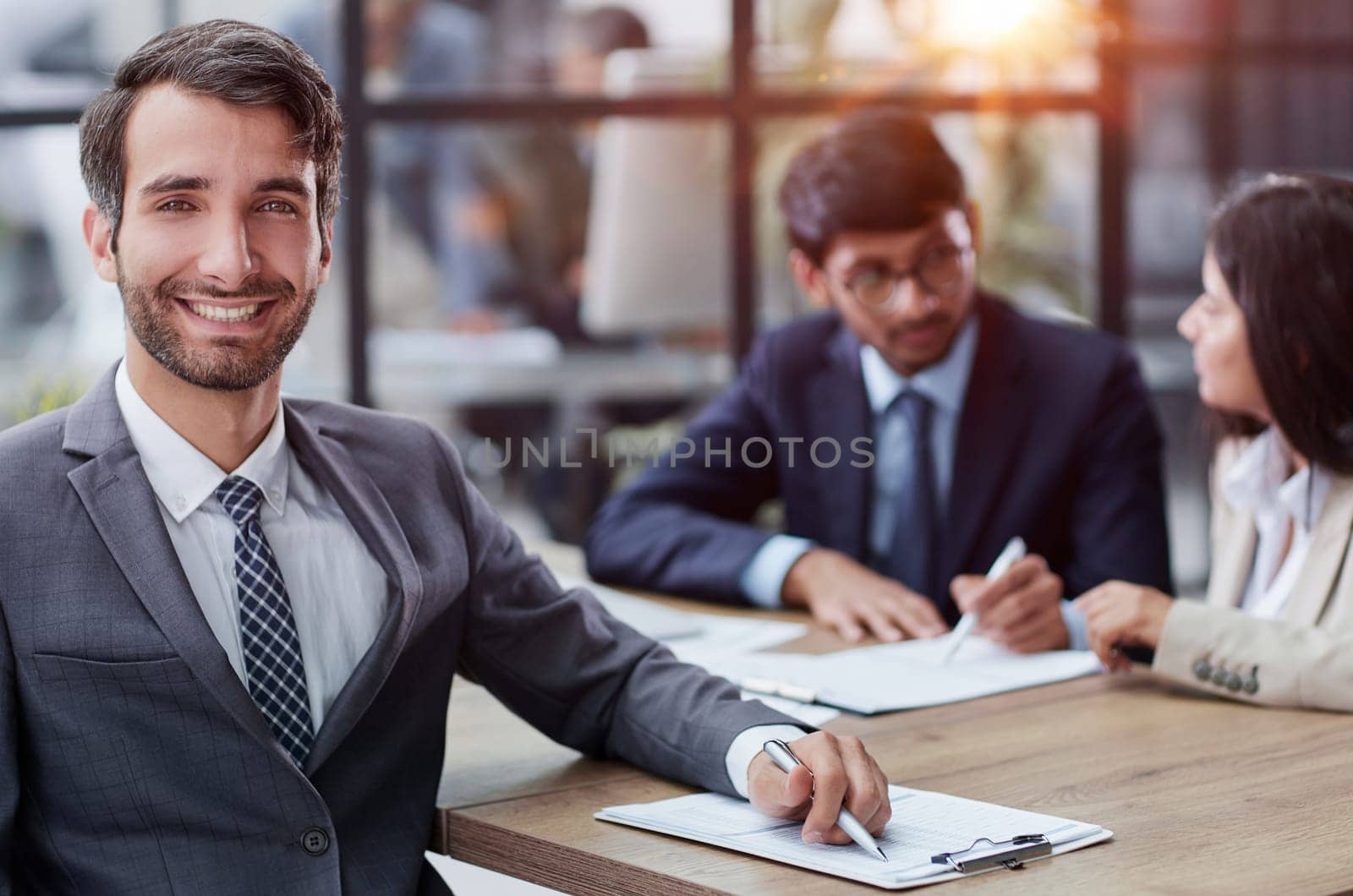young man posing for the camera while sitting at a table in the office against the background of his colleagues
