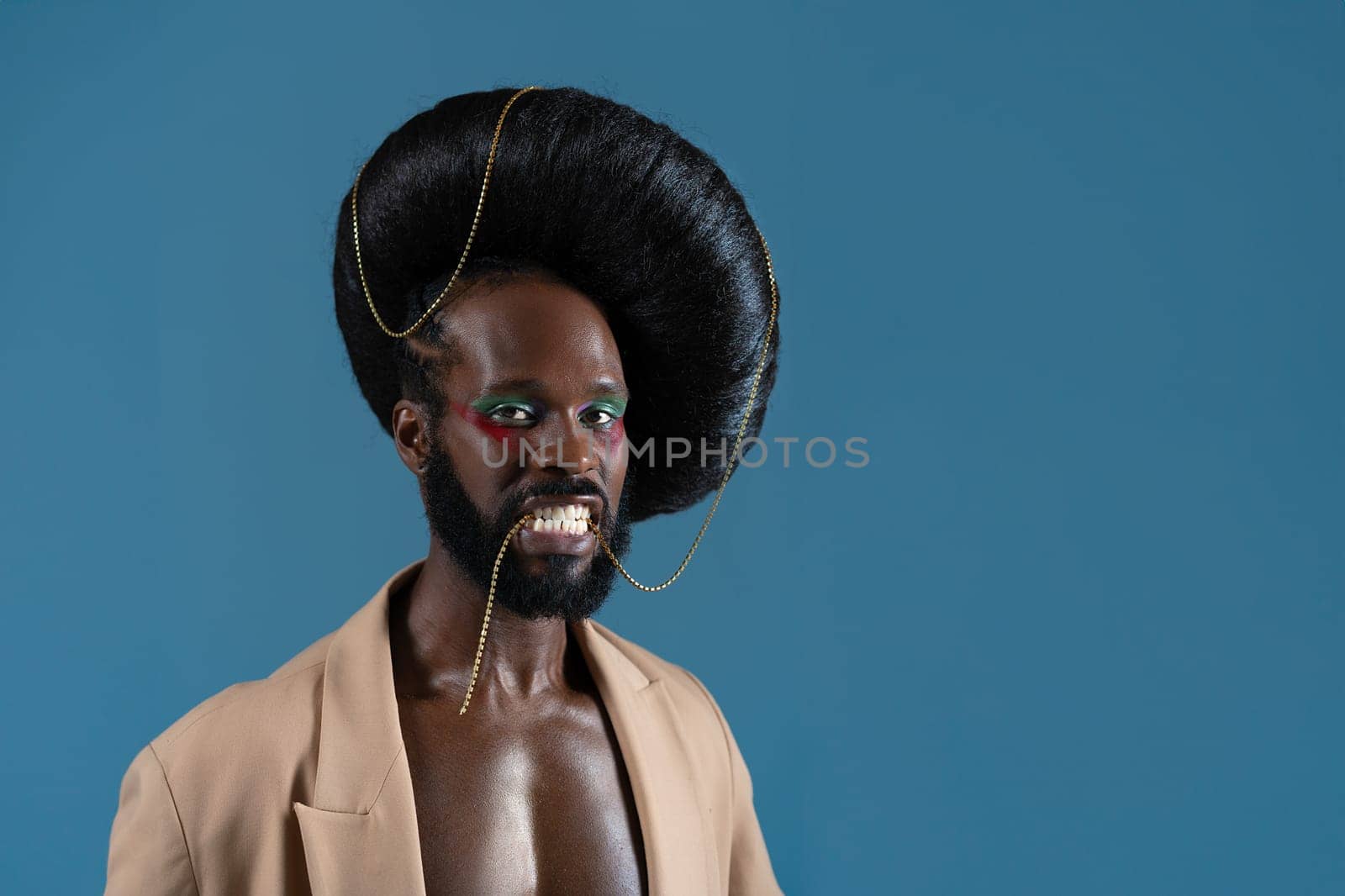 Portrait of African American gay man with makeup and black vintage wig. LGBT person holding gold accessory with teeth and wearing jacket on blue background.