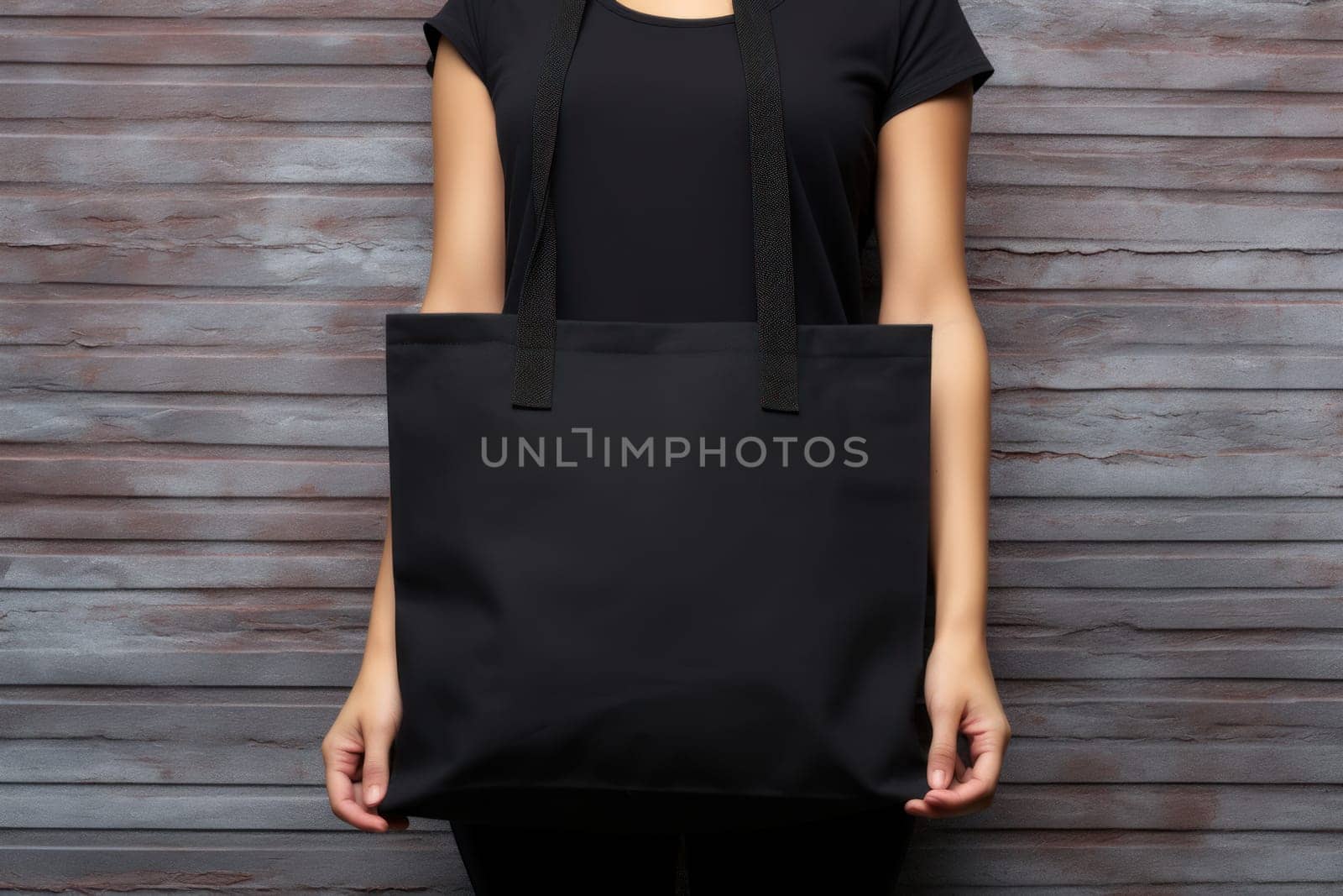 Hand holding black cotton tote bag mockup on a wall background.