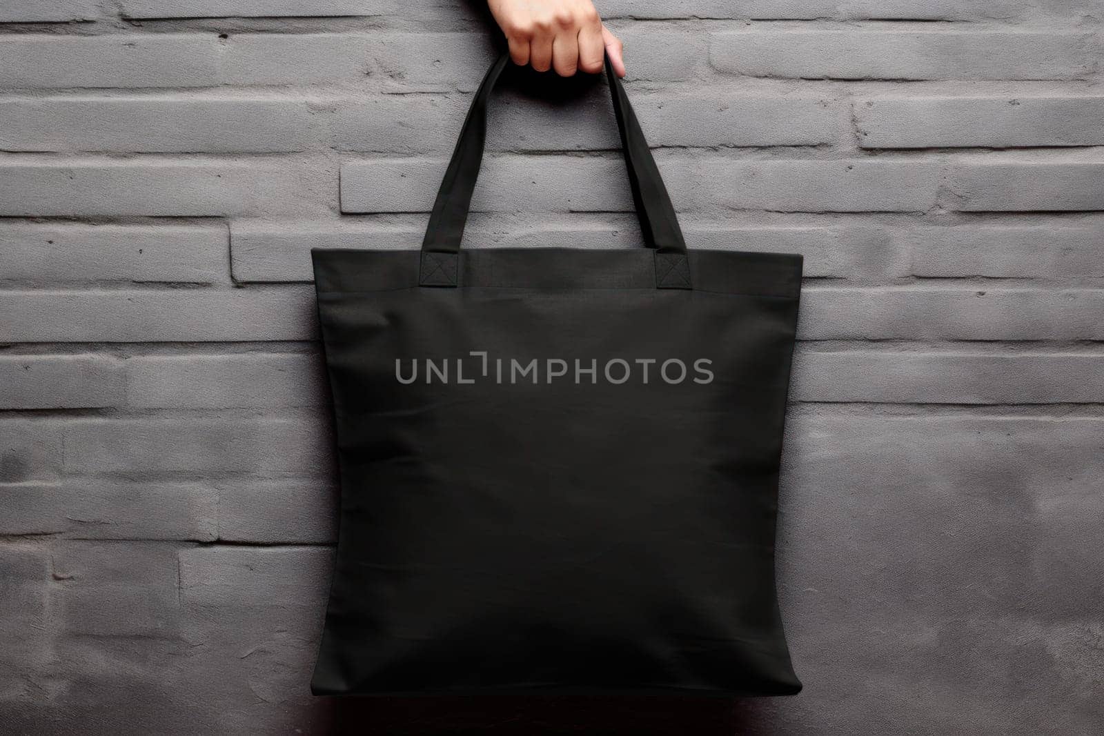 Hand holding black cotton tote bag mockup on a wall background.