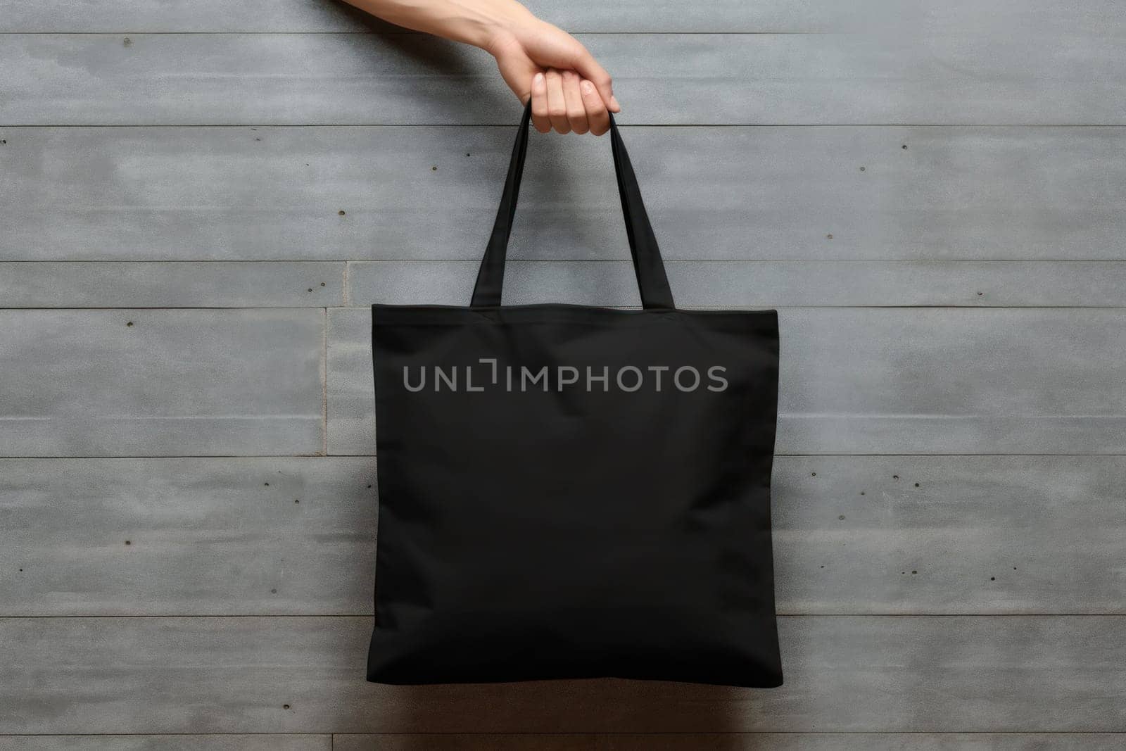 Hand holding black cotton tote bag mockup on a wall background.