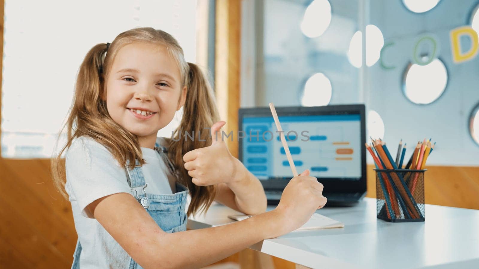 Smiling girl using laptop while look and show thumb up to camera. Cute child wearing headphone while working by using laptop writing code in STEM technology education. Online learning. Erudition.