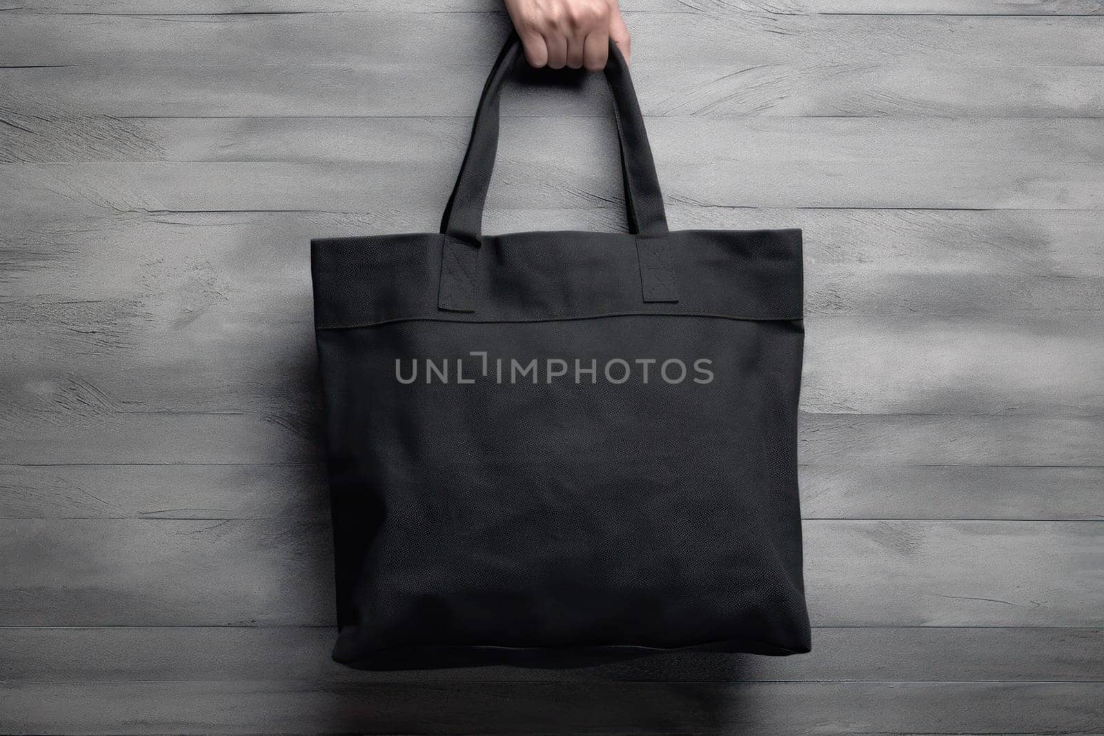 Hand holding black cotton tote bag mockup on a wall background.