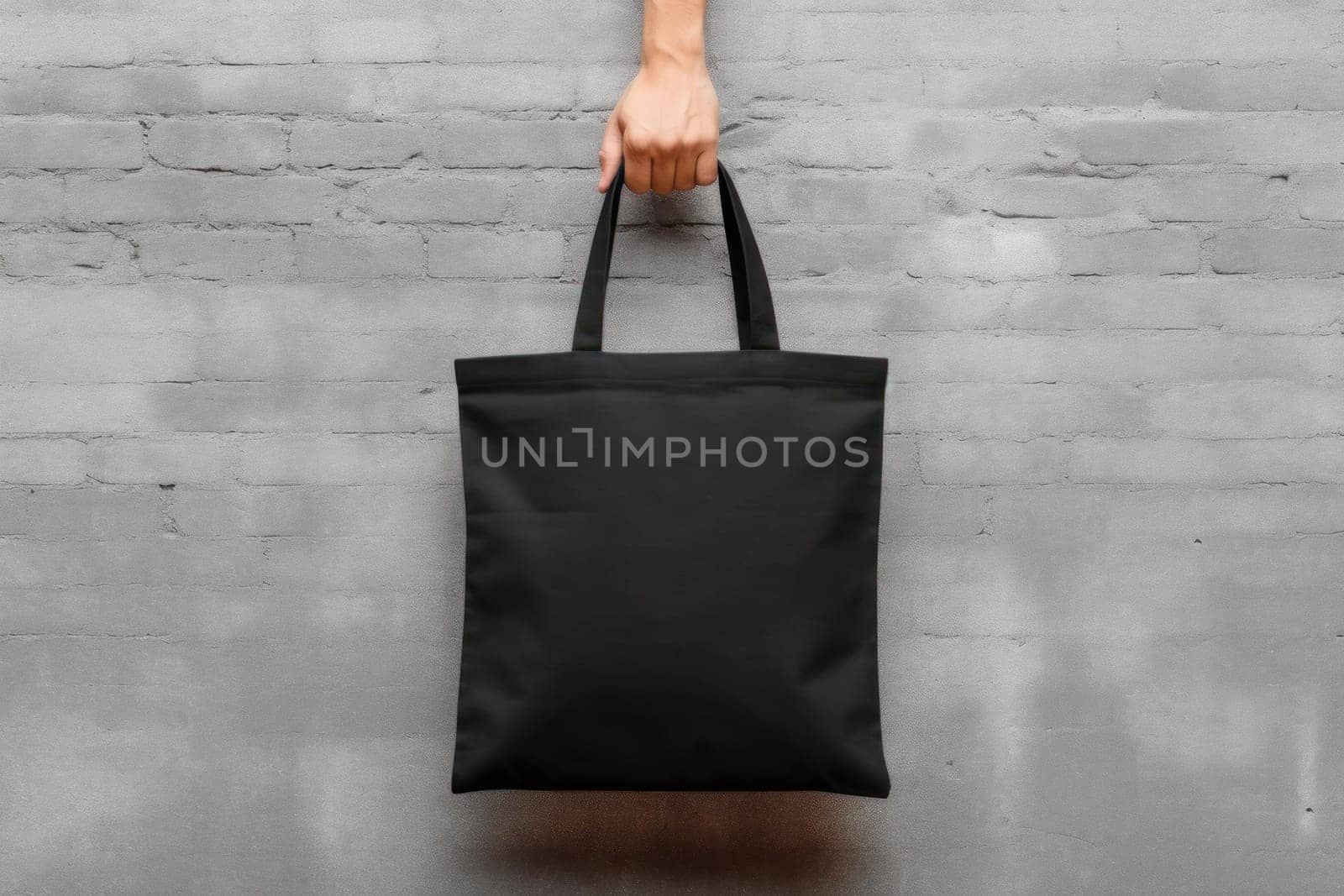 Hand holding black cotton tote bag mockup on a wall background.