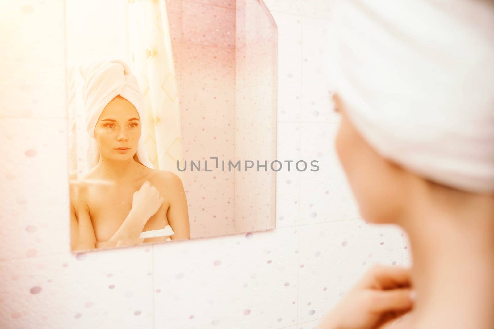 Portrait of young girl with towel on head in white bathroom looks and touches her face in the mirror and enjoys youth and hydration. Natural beauty, home care for problem skin.
