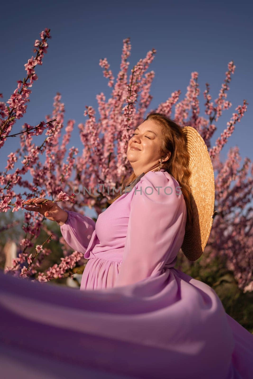 Woman blooming peach orchard. Against the backdrop of a picturesque peach orchard, a woman in a long pink dress and hat enjoys a peaceful walk in the park, surrounded by the beauty of nature. by Matiunina