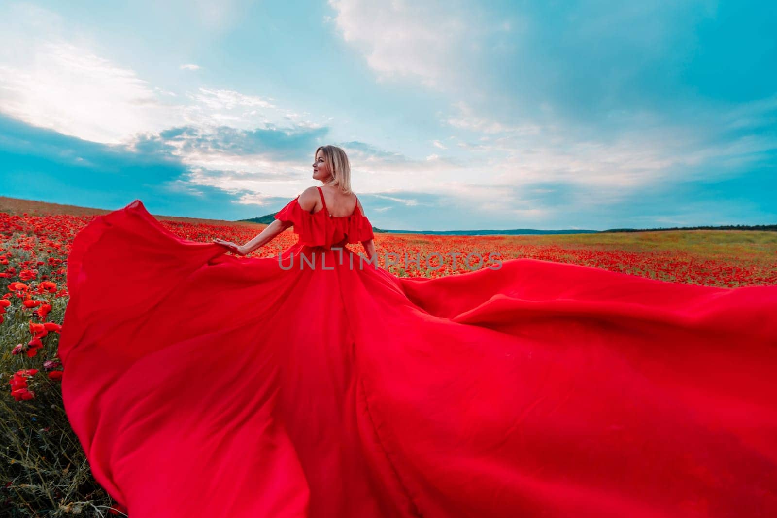 Woman poppy field red dress. Happy woman in a long red dress in a beautiful large poppy field. Blond stands with her back posing on a large field of red poppie