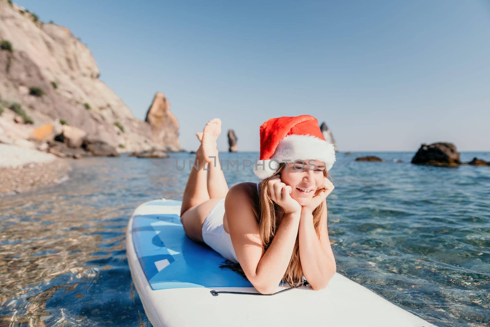 Woman sea sup. Close up portrait of happy young caucasian woman with long hair in Santa hat looking at camera and smiling. Cute woman portrait in a white bikini posing on sup board in the sea by panophotograph