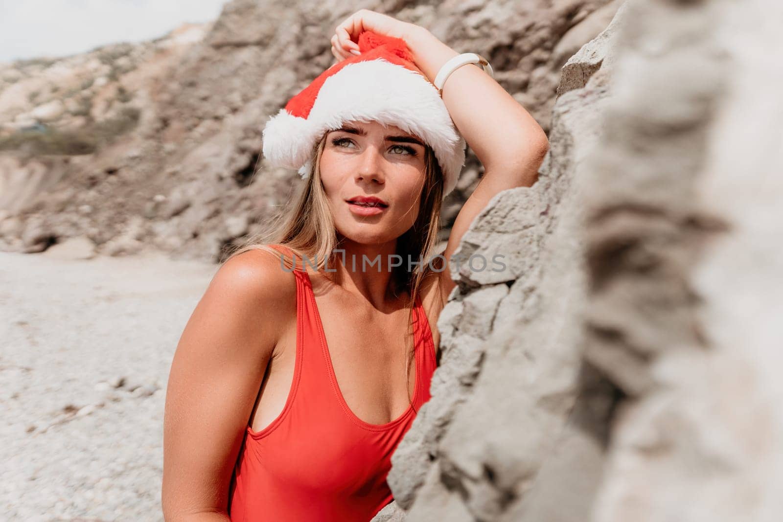 Woman summer travel sea. Happy tourist in red bikini and Santas hat enjoy taking picture outdoors for memories. Woman traveler posing on the beach surrounded by volcanic mountains, sharing travel joy by panophotograph