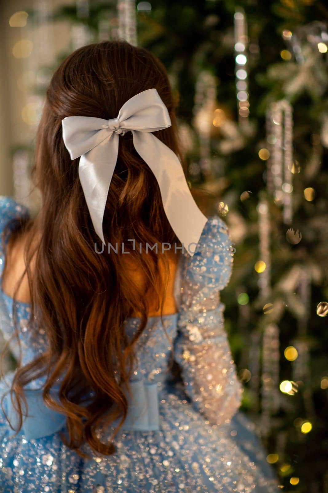 The girl decorates the Christmas tree, holding Christmas baubles in her hand. She wears a blue dress with a bow in her hair. Holiday concept. by Matiunina