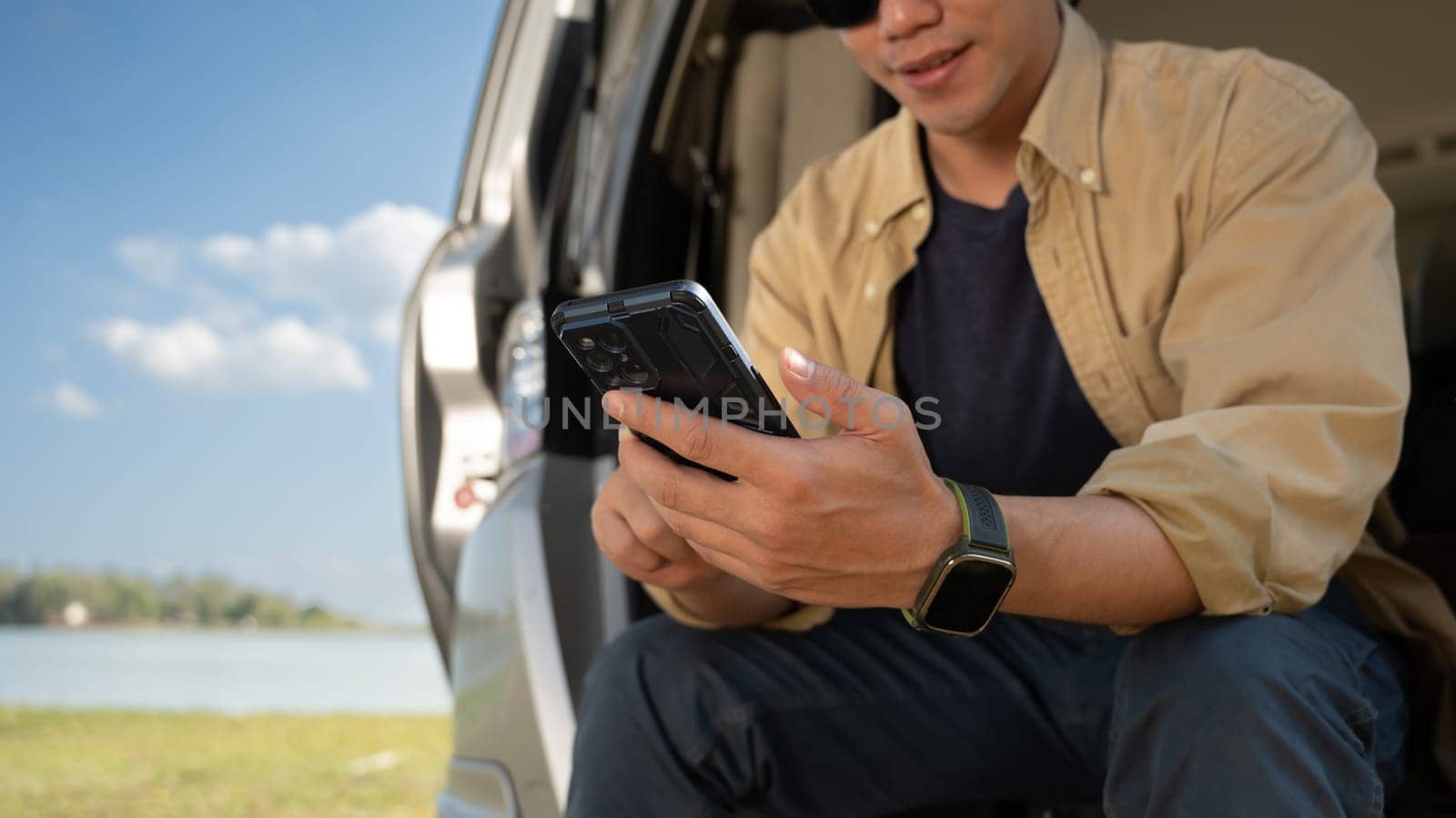 Young solo traveler sitting on the open trunk and using mobile phone over blue sky. Travel lifestyle concept.