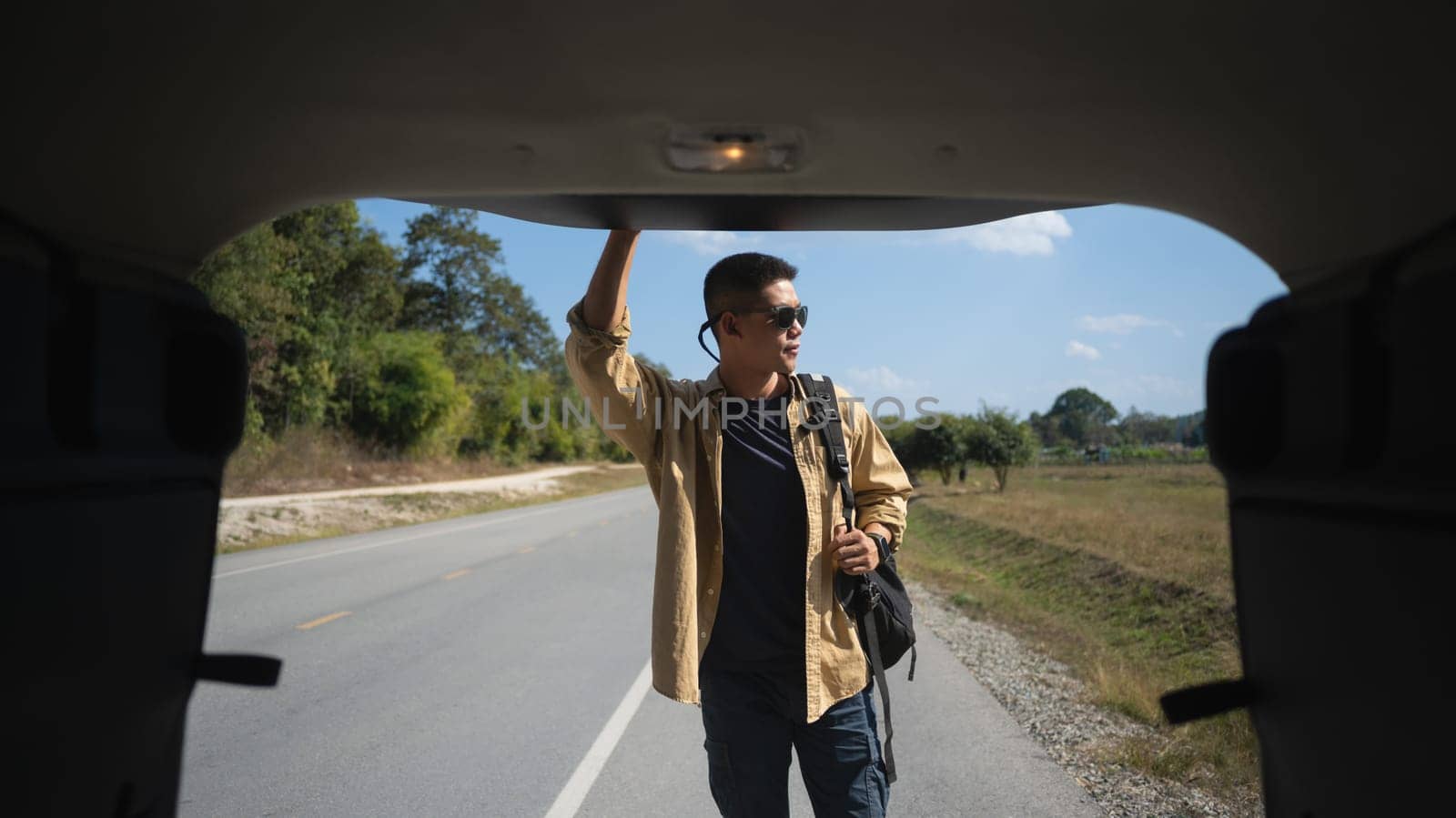 Solo travel man carrying backpack standing on countryside road a seen through open vehicle trunk.