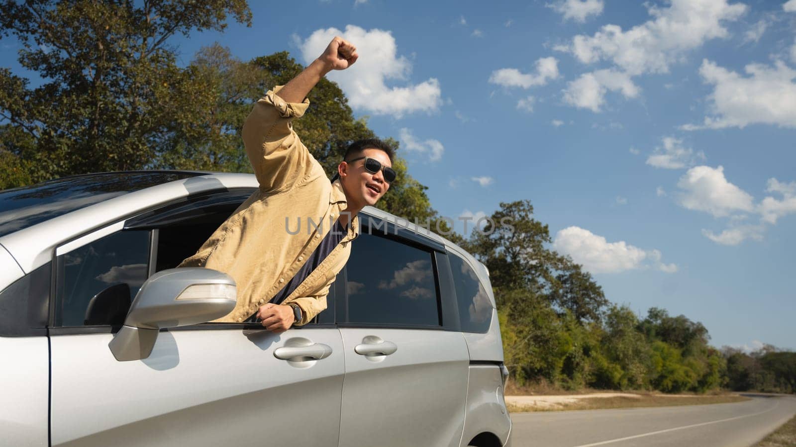 Happy man leaning out of car window, enjoying nature on country road. Travel and active lifestyle concept.
