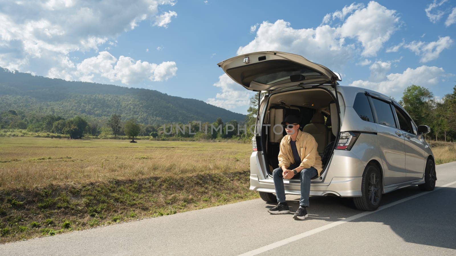 Young male solo traveler sitting on the open trunk of car with beautiful mountain views and blue sky.