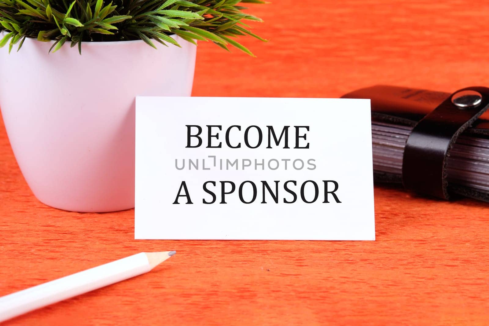 BECOME A SPONSOR text written on a business card next to a business card holder, a green flower and a pencil on an orange background