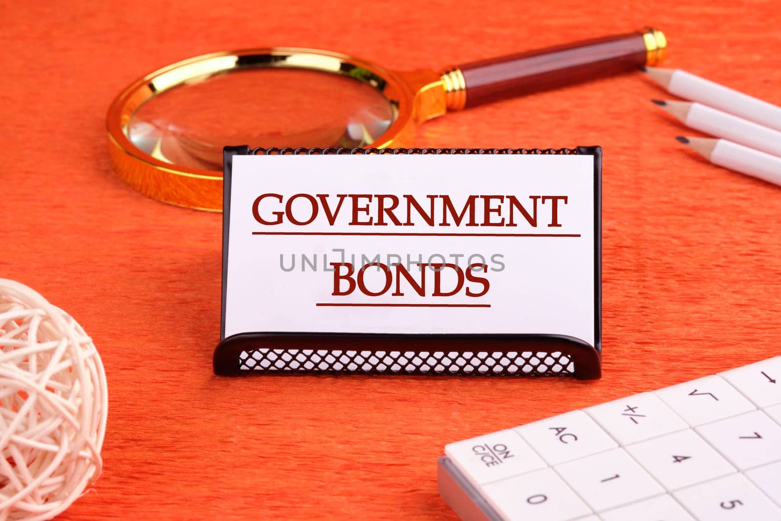 Government Bonds text on a white business card on a stand on an orange background, next to a magnifying glass, a calculator and pencils