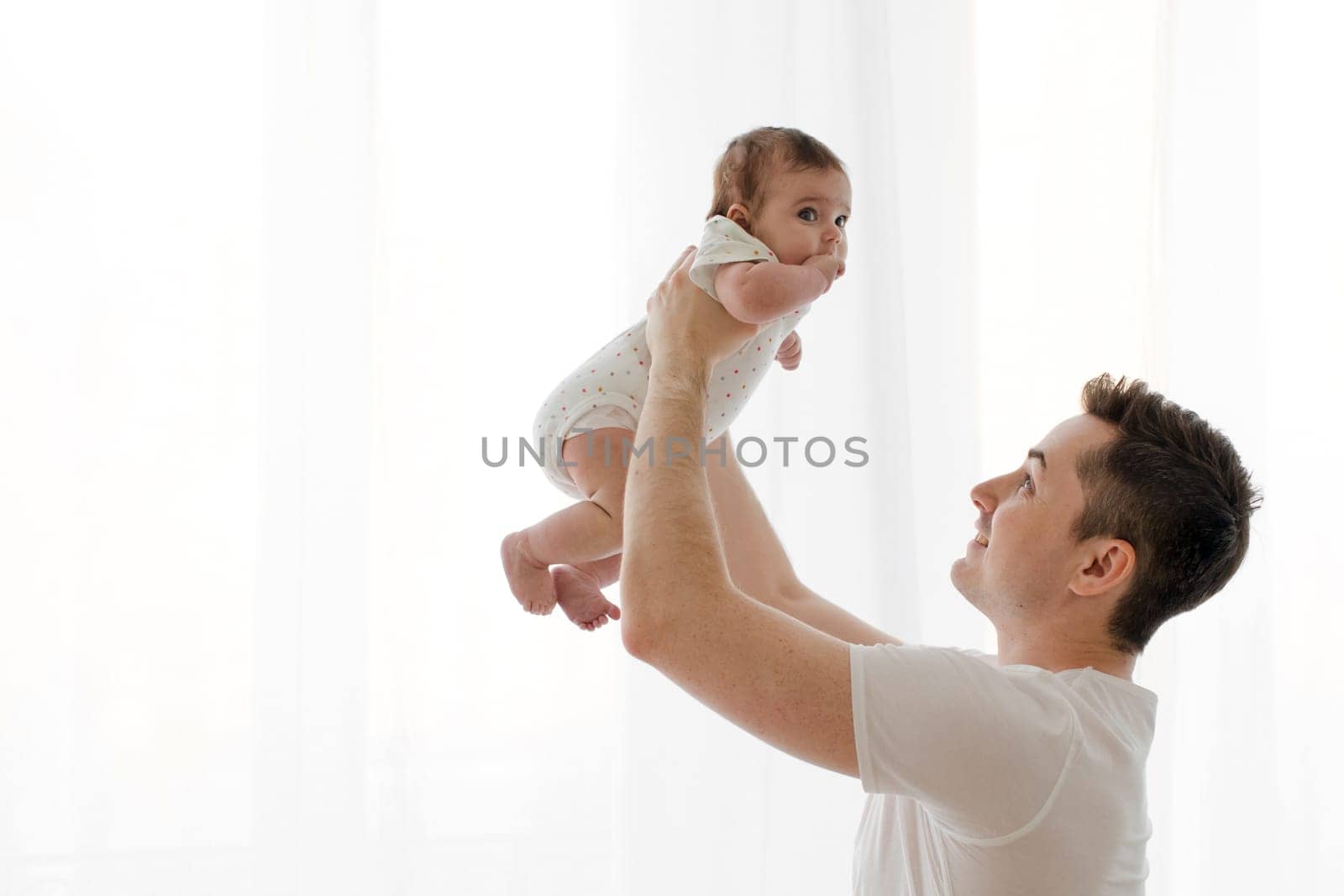 Side view of happy young dad in white t shirt lifting in air and playing with cute newborn child while bonding at home against white background