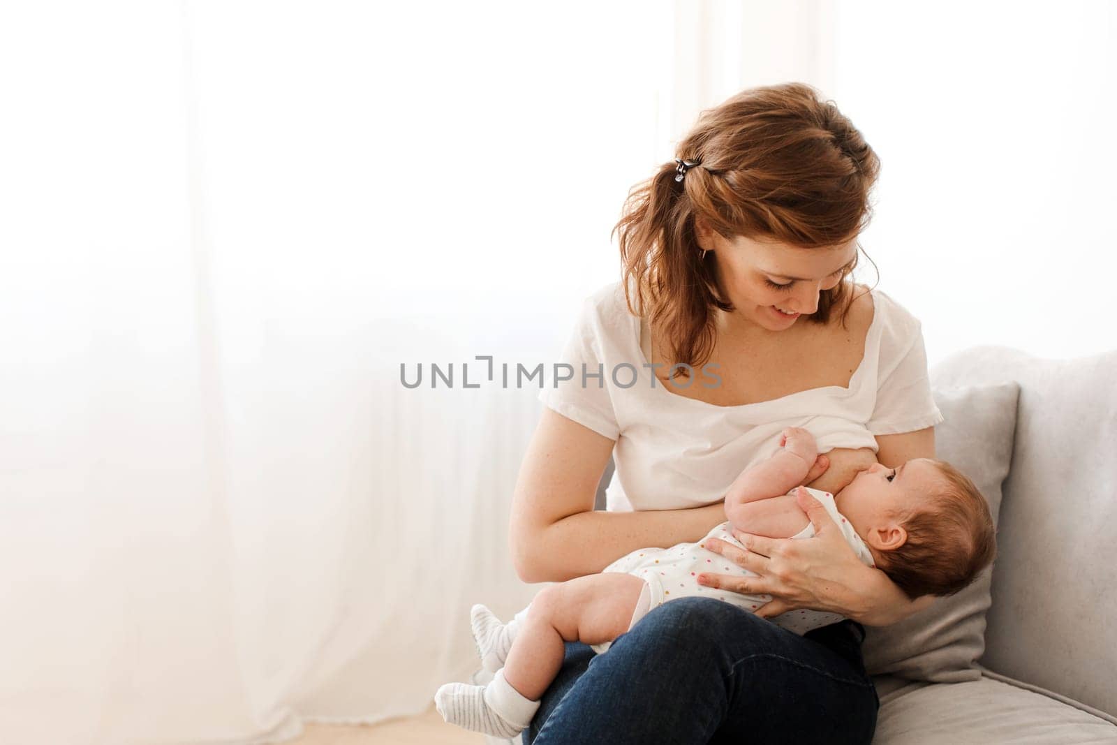 Happy brunette woman wearing cream t shirt smiling and sitting on couch while nursing and breastfeeding adorable infant child in light living room
