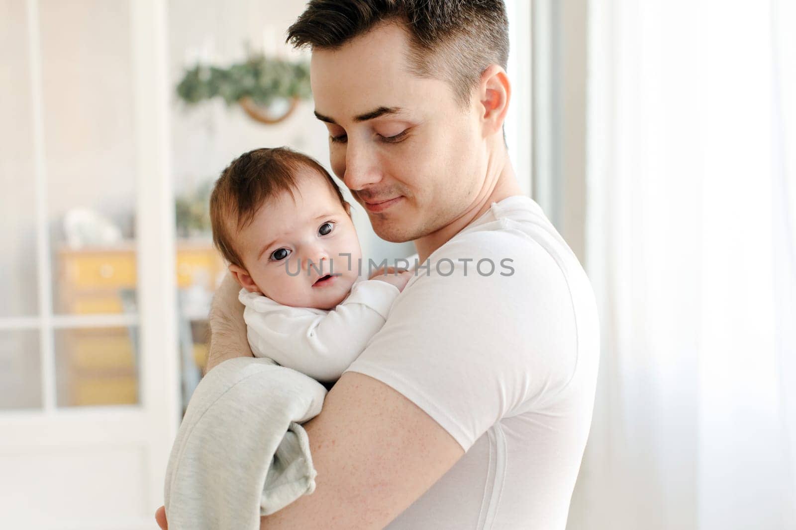 Side view of smiling young crop father in casual clothes looking down while embracing cute little baby looking at camera and standing outside in daylight against blurred background