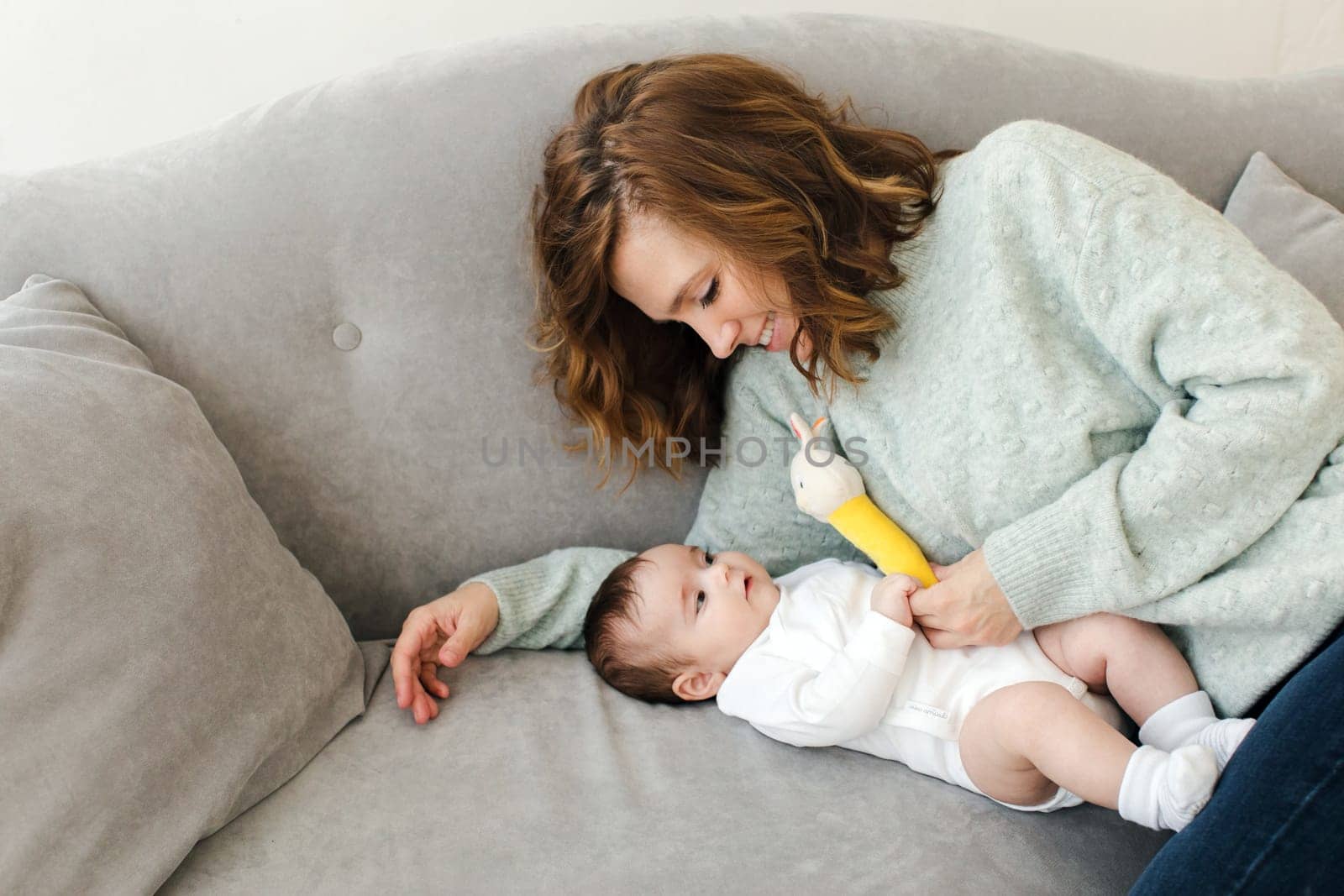 High angle of smiling young mother and cute child looking at each other while lying on sofa in daylight and playing with bunny toy in living room