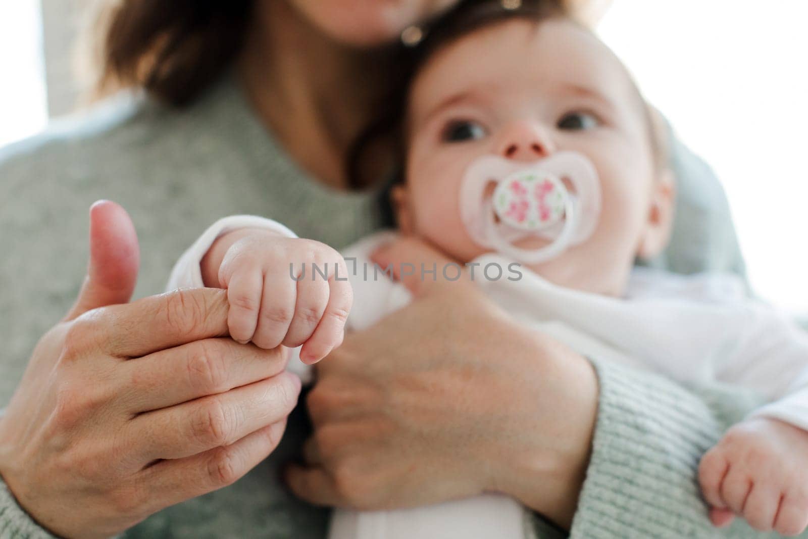 Unrecognizable caring mom in sweater hugging and touching hand of cute baby sucking pacifier while bonding at home blurred background