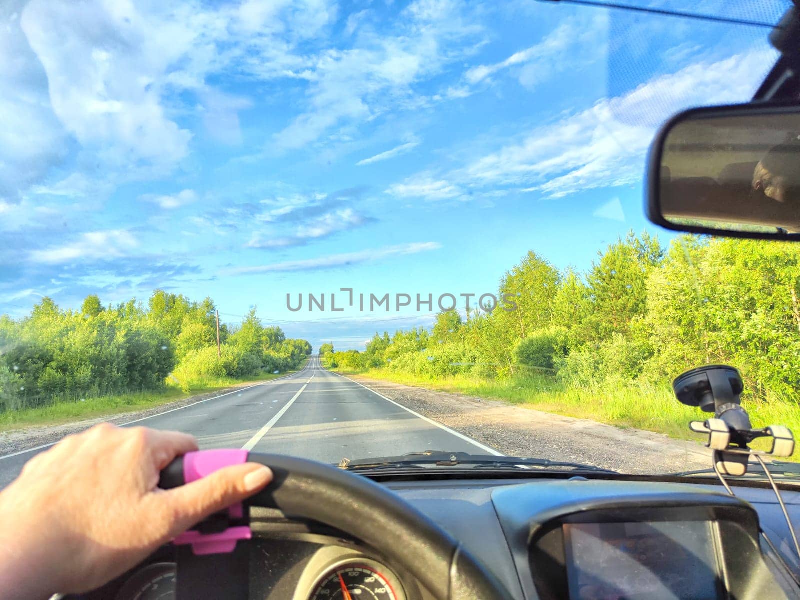 Car salon, windshield, hand of woman on steering wheel and landscape. View from seat of driver on nature with Road, trees, blue sky at a sunny day. Single trip of female traveler. Partial focus